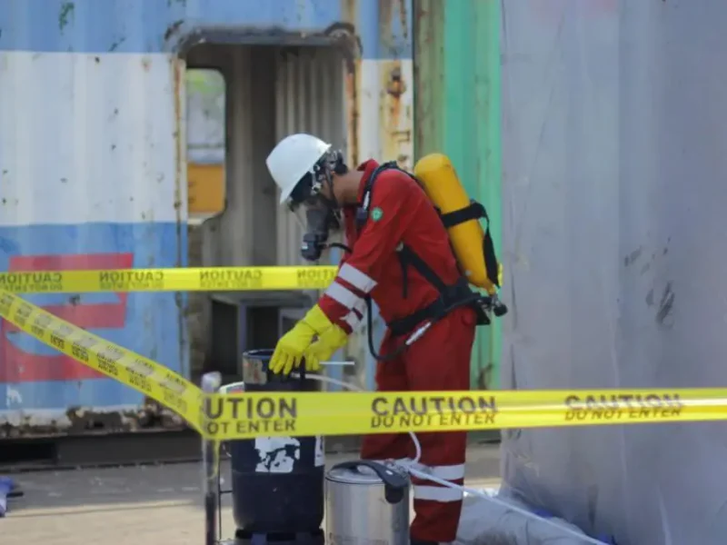 Worker in red protective suit and gas mask handling equipment behind yellow caution tape
